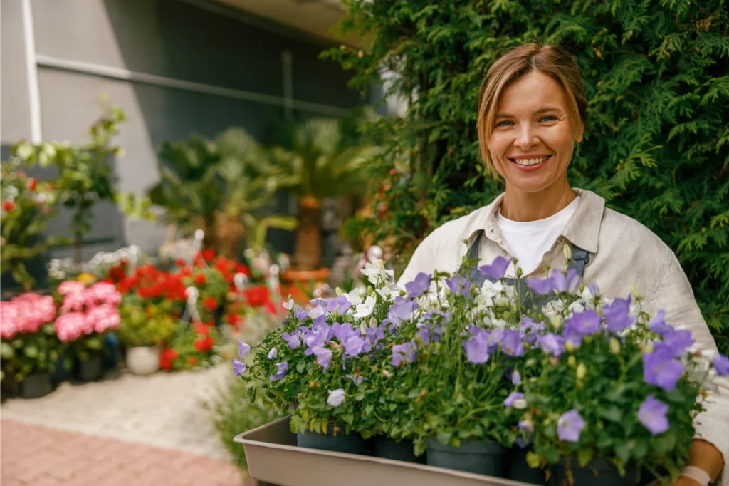 Smiling plant vendor at outdoor garden market with purple blooms in large planter tray